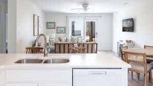 A modern kitchen with a central island, dining area with wicker chairs, and a view of the sea through a glass door. - Seabatical East, Almost Oceanfront Duplex in Emerald Isle, NC! (Emerald Isle)