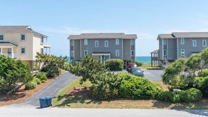 A coastal residential area with three two-story buildings, a paved driveway, and a parking area with cars. - Seabatical East, Almost Oceanfront Duplex in Emerald Isle, NC! (Emerald Isle)