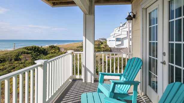 Ein überdachter Balkon mit zwei türkisfarbenen Stühlen, einem weißen Geländer und Blick auf Strand und Meer.