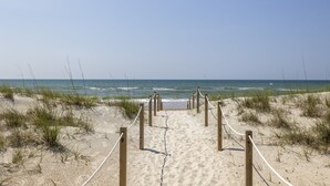 Ein sandiger Küstenpfad führt zum Meer, mit Holzpfählen und Seilen als Wegweiser.