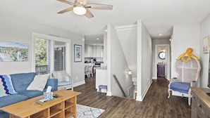 Ein modernes Wohnzimmer mit einer Couch, einem Couchtisch und einem Deckenventilator. Eine Treppe führt zu einer weiteren Ebene, und im Hintergrund ist eine Küchenzeile erkennbar.