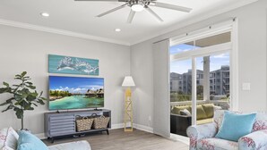 A modern living room with a flat-screen TV, a ceiling fan, and a large window with a view of buildings. - Seaside Villas 1, Almost Oceanfront Duplex in Atlantic Beach, NC! (Atlantic Beach)