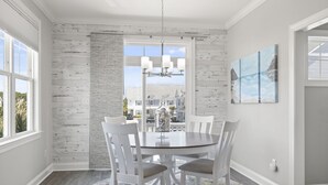 A dining room with a round table, white chairs, a chandelier, and a view of houses through large windows. - Seaside Villas 1, Almost Oceanfront Duplex in Atlantic Beach, NC! (Atlantic Beach)