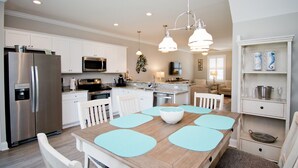 A modern kitchen with white cabinetry, stainless steel appliances, and a dining area with a wooden table and blue placemats. - Seaside Villas 44, Soundside Townhouse in Atlantic Beach, NC! (Atlantic Beach)