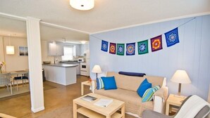 A modern living room with a sofa, coffee table, and lamps. There is a kitchen in the background and a dining area with a table and chairs. - Sunny Daze, Almost Oceanfront Duplex in Atlantic Beach, NC! (Atlantic Beach)