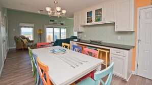 Une cuisine lumineuse avec des meubles blancs, une table à manger entourée de chaises colorées et une vue sur l’océan par la fenêtre.