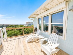 Une terrasse en bois avec des fauteuils Adirondack blancs, de grandes fenêtres et une vue sur une maison et la verdure.
