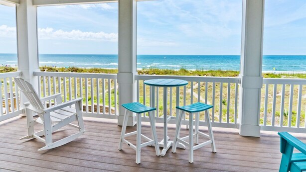 A covered outdoor area with a wooden deck, white railings, a white rocking chair, and two blue bar stools. - Swiss House, Oceanfront Home in Atlantic Beach, NC! (Atlantic Beach)