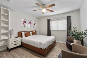 Ein Schlafzimmer mit einem Bett, Nachttischen, einem Deckenventilator, einem Fenster mit Vorhängen und einer Pflanze.