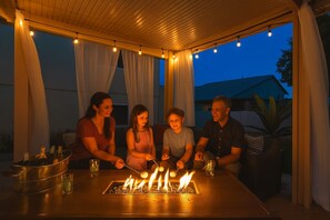 A group of people around a table with a fire feature, under a covered area with string lights. - Epic 6BR w/ Foosball + Popcorn | Pool + Fire Pit (Peoria)
