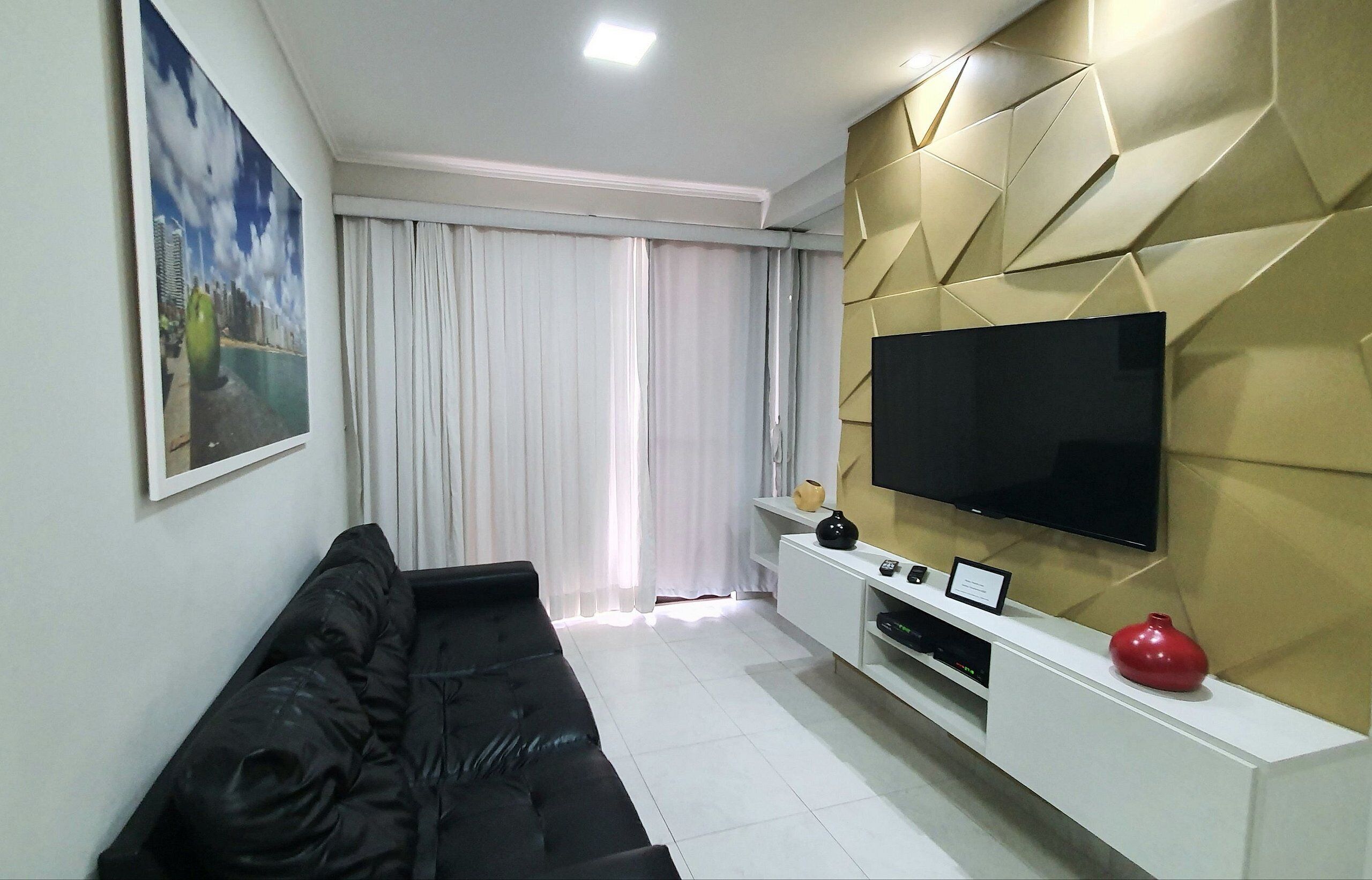 A modern living room with a black sofa, a flat-screen TV mounted on a wall with geometric panels, a white entertainment unit, and a large window with white curtains.