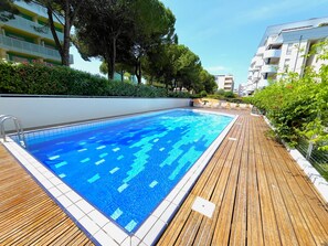 Una piscina con bordo in legno, circondata da vegetazione e edifici.