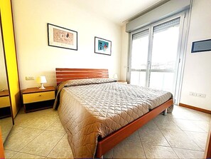 Um quarto com uma cama, mesa de cabeceira, abajur e duas imagens emolduradas na parede.
