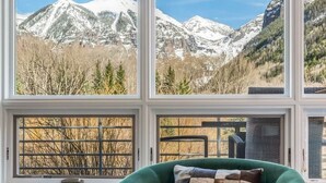 A room with a green armchair, a table with a book and remote, and a window with a mountain view.