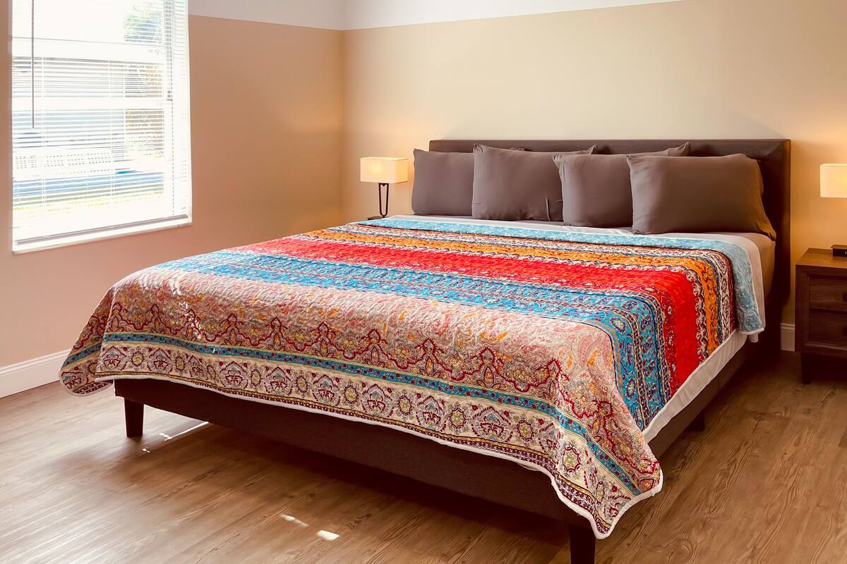 A bedroom with a bed, two bedside tables, a window with blinds, and a patterned bedspread.