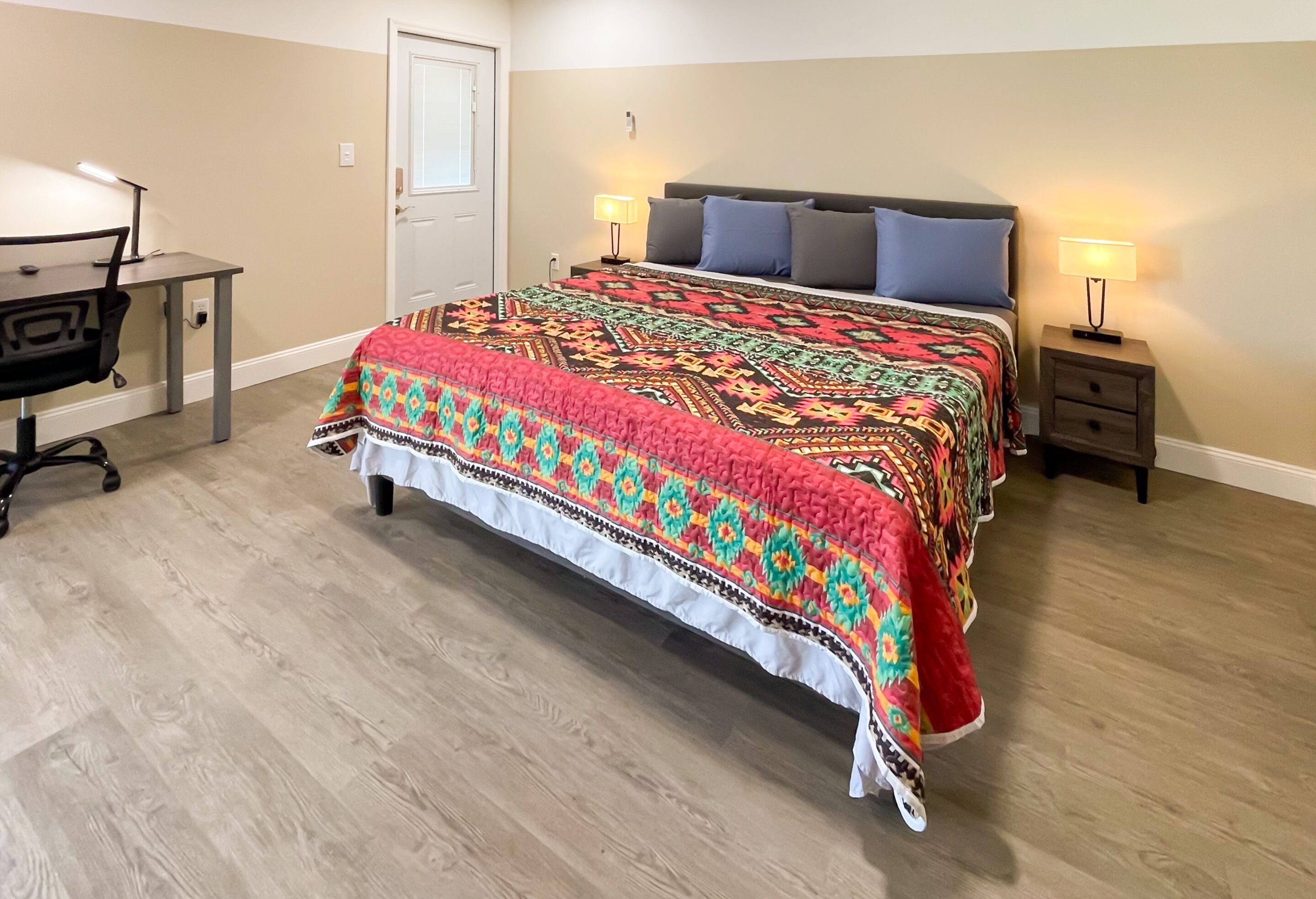 A bedroom with a bed, two bedside tables with lamps, a desk with a chair, and a door.