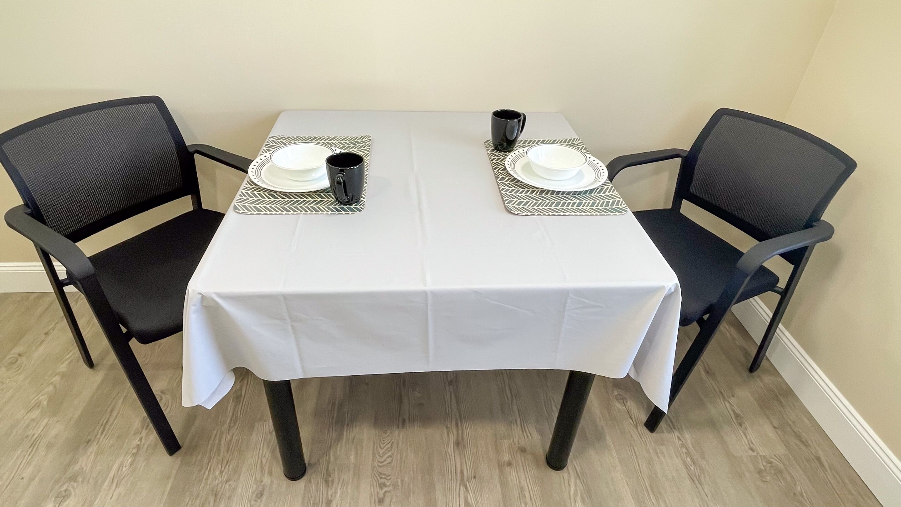 A dining table with a white tablecloth, two black chairs, and two place settings with plates, bowls, and black mugs.