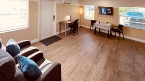 A modern hotel room with a wooden floor, a sofa with blue cushions, a desk with a chair, a TV mounted on the wall, and a window with blinds.