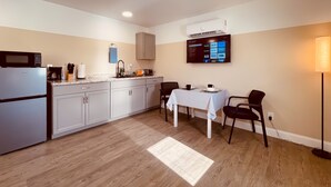 A compact kitchen with a refrigerator, microwave, and a coffee maker. There's a dining area with a table and chairs, and a flat-screen TV mounted on the wall displaying various channels.