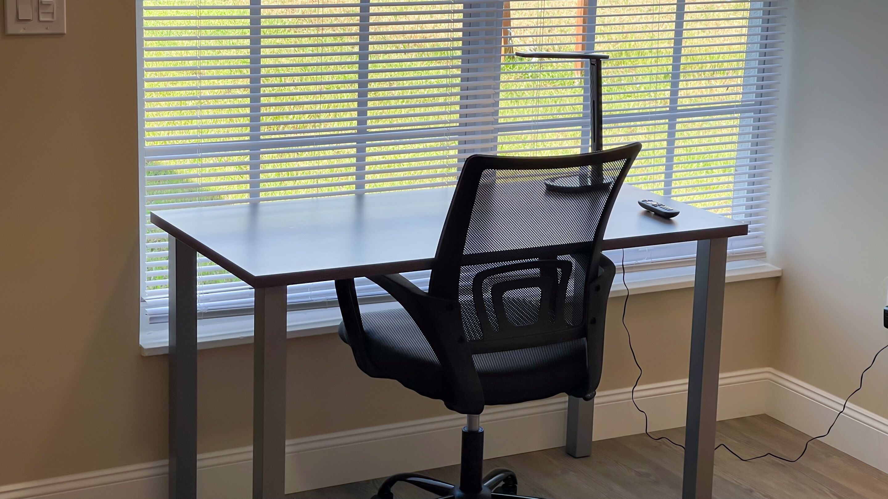 Un espace de travail comprenant un bureau et une chaise de bureau à dossier en maille, placés près d’une fenêtre munie de stores.