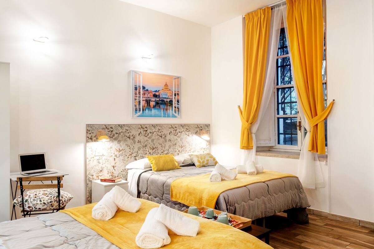 A hotel room with two beds, a desk, and a window with yellow curtains.