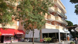 A building with a red awning labeled "MACELLERIA" and "METZGEREI" on the ground floor, a white awning with "GARDEN" on the second floor, and a tree in the foreground. - Nice Flat for 6 Guests in Bibione Pineda (San Michele al Tagliamento)