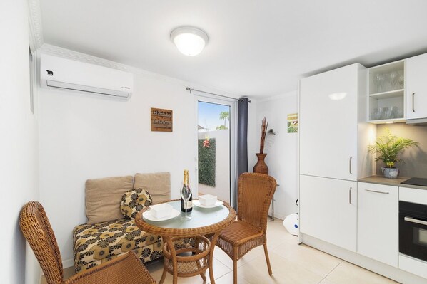 A compact living space with a kitchenette, a dining area with wicker furniture, a sofa with patterned cushions, and a door leading to an outdoor area.