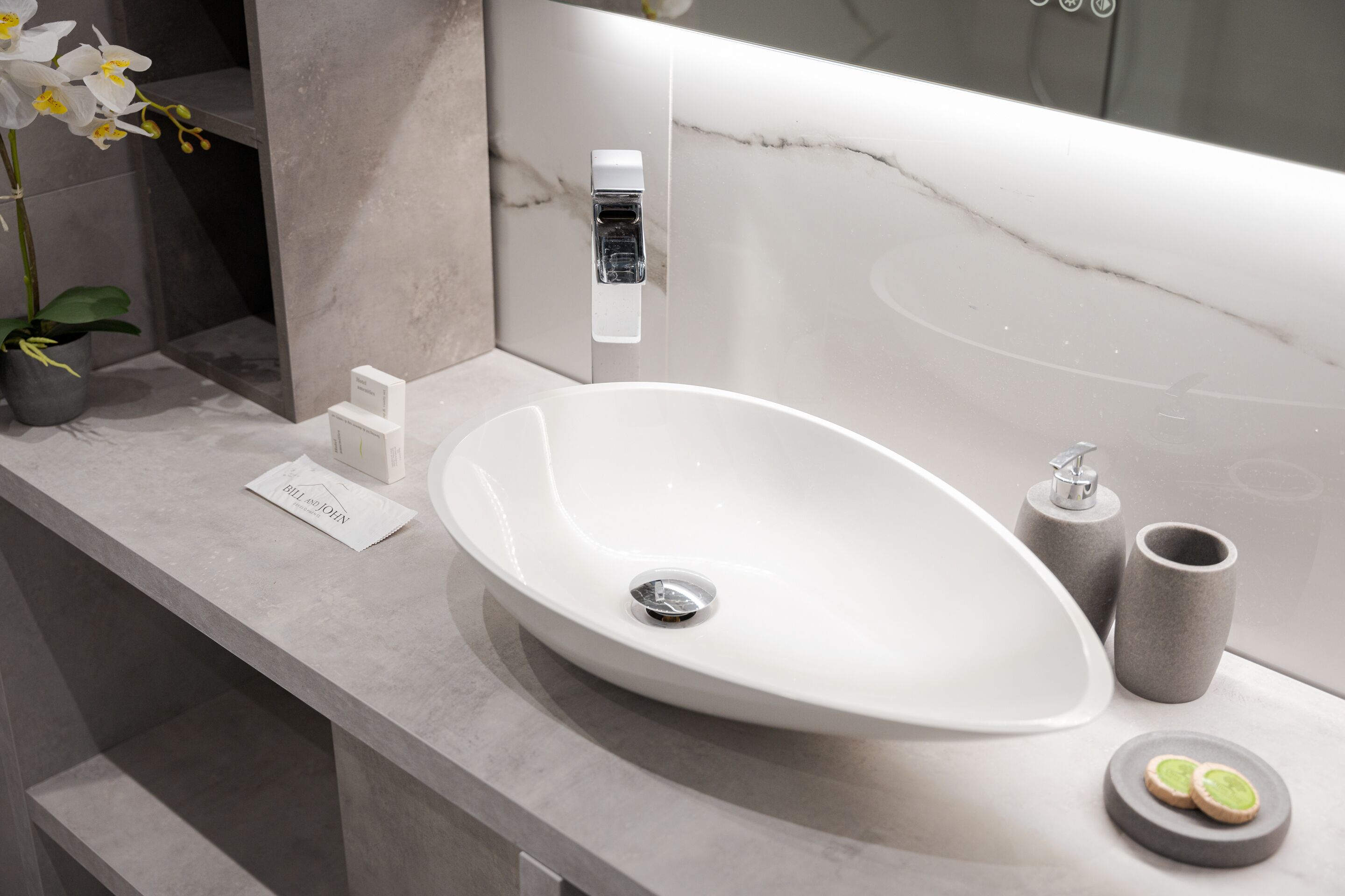 A modern bathroom with a large oval sink, a marble backsplash, a potted orchid, and a small sign.