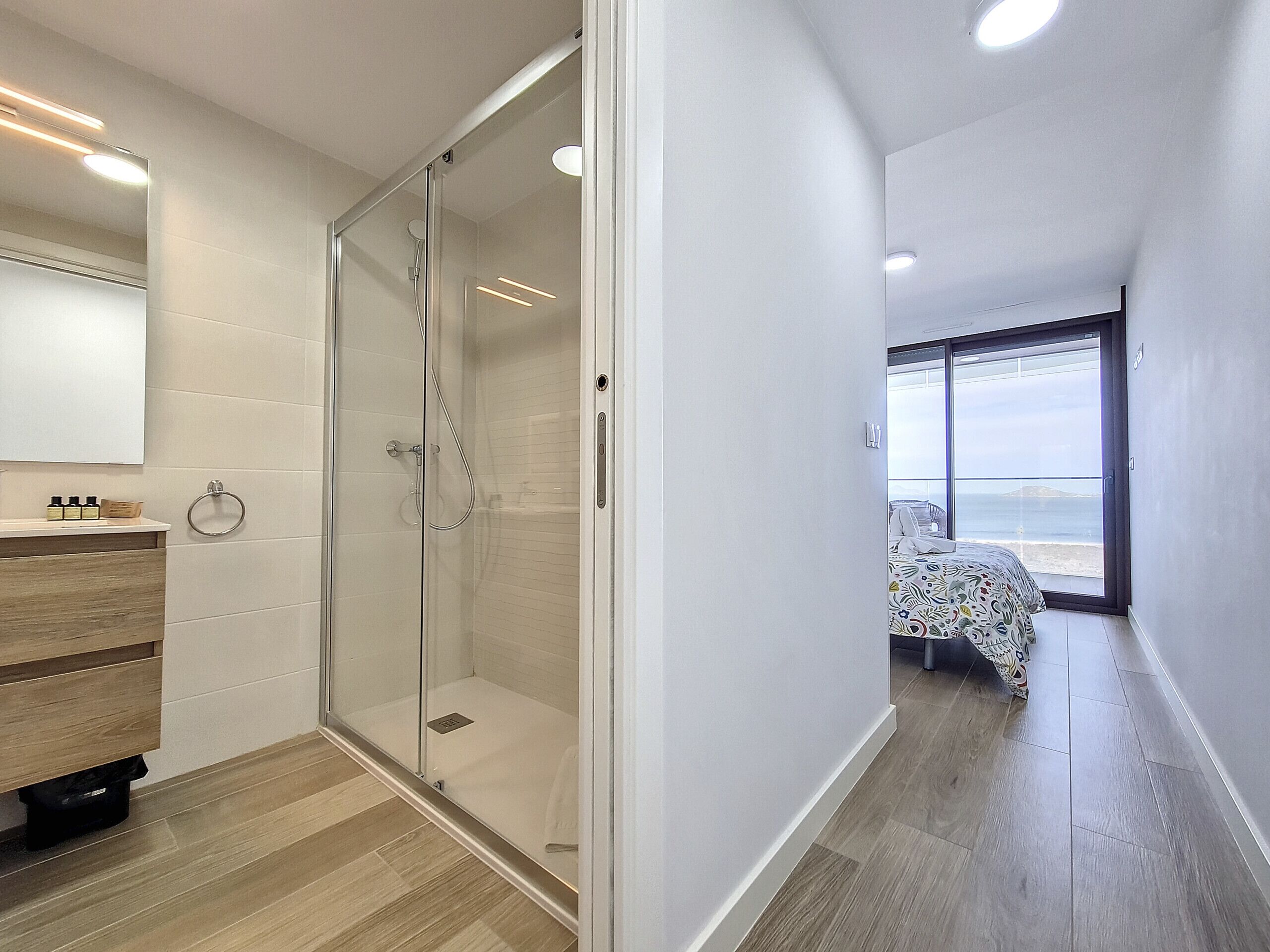A modern bathroom with a glass-enclosed shower, a wooden vanity, and a large window offering a view of the sea.