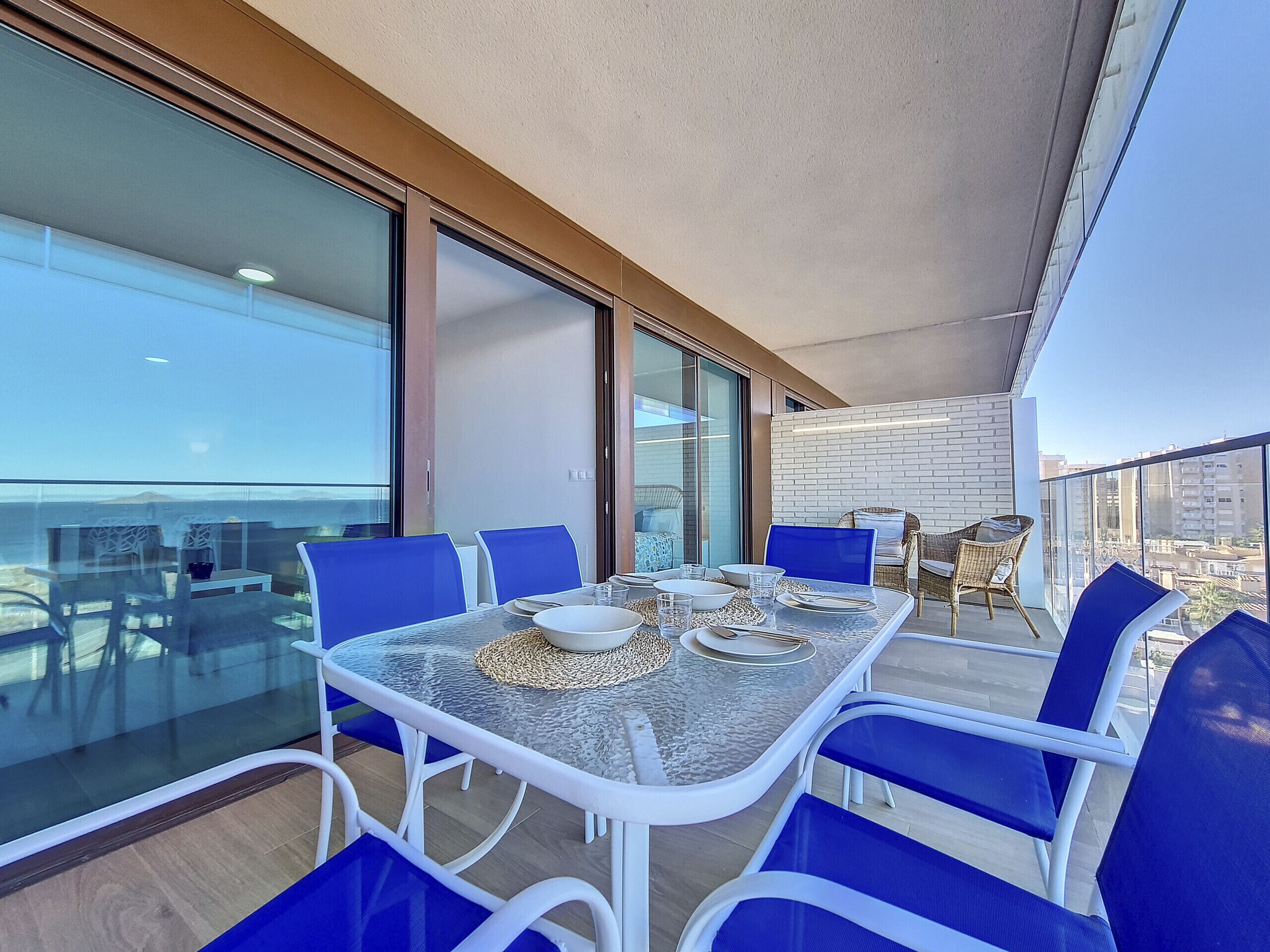Een modern balkon met een glazen eettafel, blauwe stoelen en een rieten stoel.