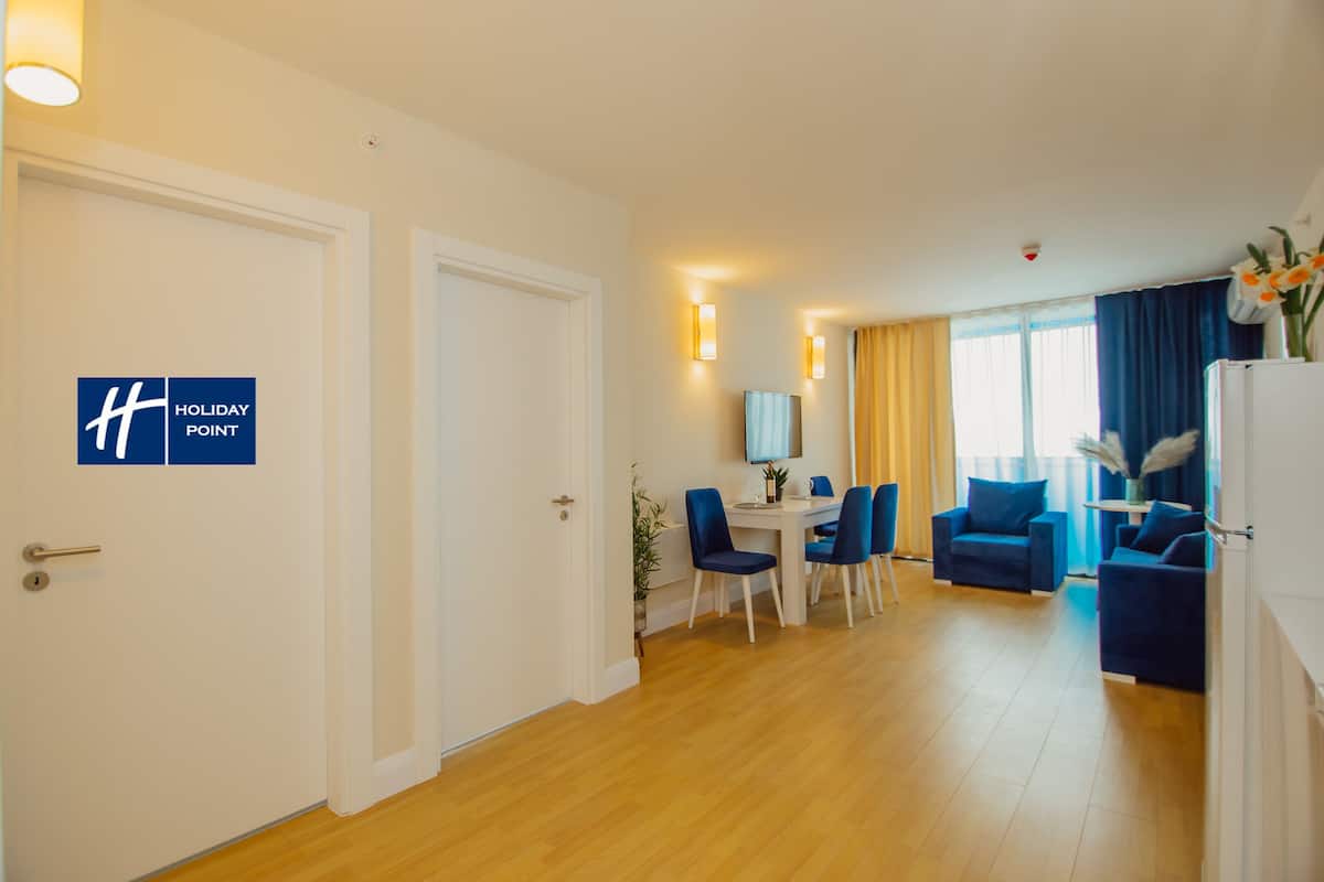 A hotel room with a dining area, a living room with blue furniture, and a sign indicating it's a Holiday Point hotel.