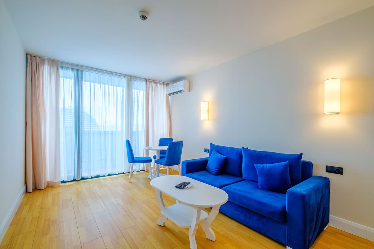 A modern living room with a blue sofa, white coffee table, and blue chairs. There is a window with sheer curtains and an air conditioning unit on the wall.