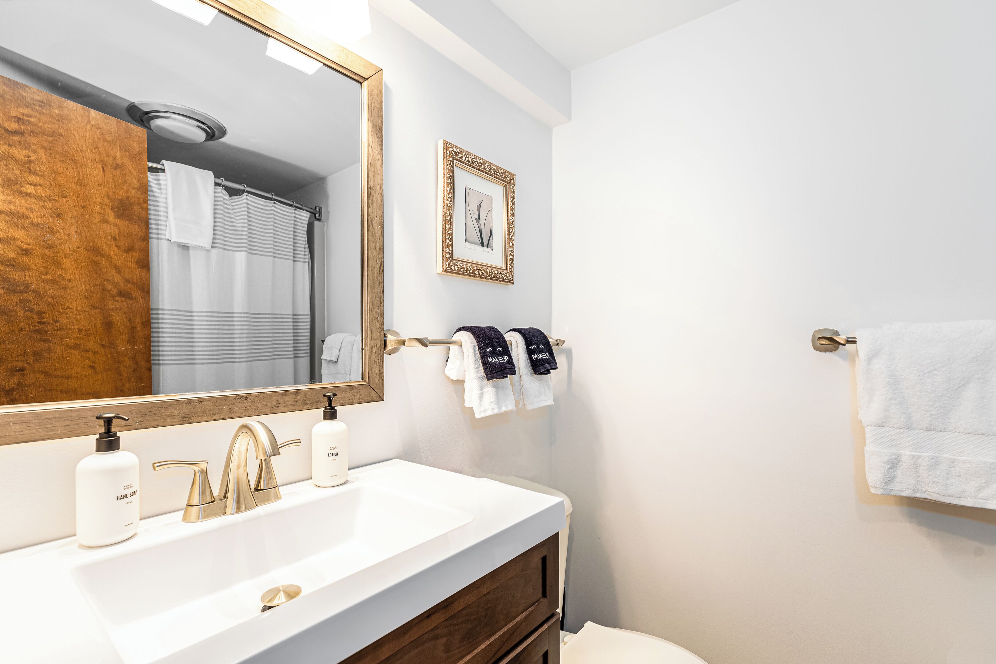A bathroom with a white sink, gold faucets, and a large mirror. There are two towels hanging on a towel rack, one white and one with "MANUEL" on it.