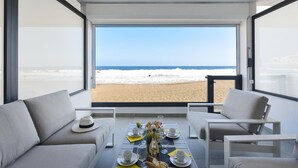 A modern room with a large glass window overlooking a beach. There is a dining table set with plates, glasses, and a bottle. - La Terraza de Salinetas By VillaGranCanaria (Telde)