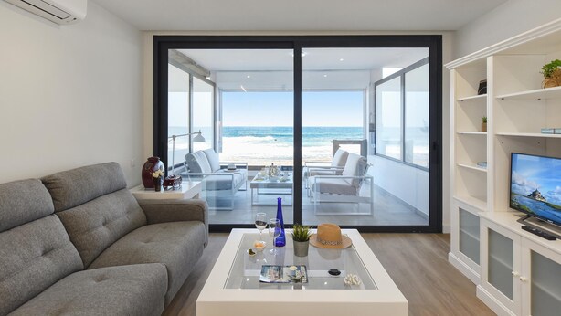 A modern living room with a grey sofa, a glass coffee table, and a TV on a white cabinet. A large sliding glass door leads to a balcony with a view of the beach. - La Terraza de Salinetas By VillaGranCanaria (Telde)