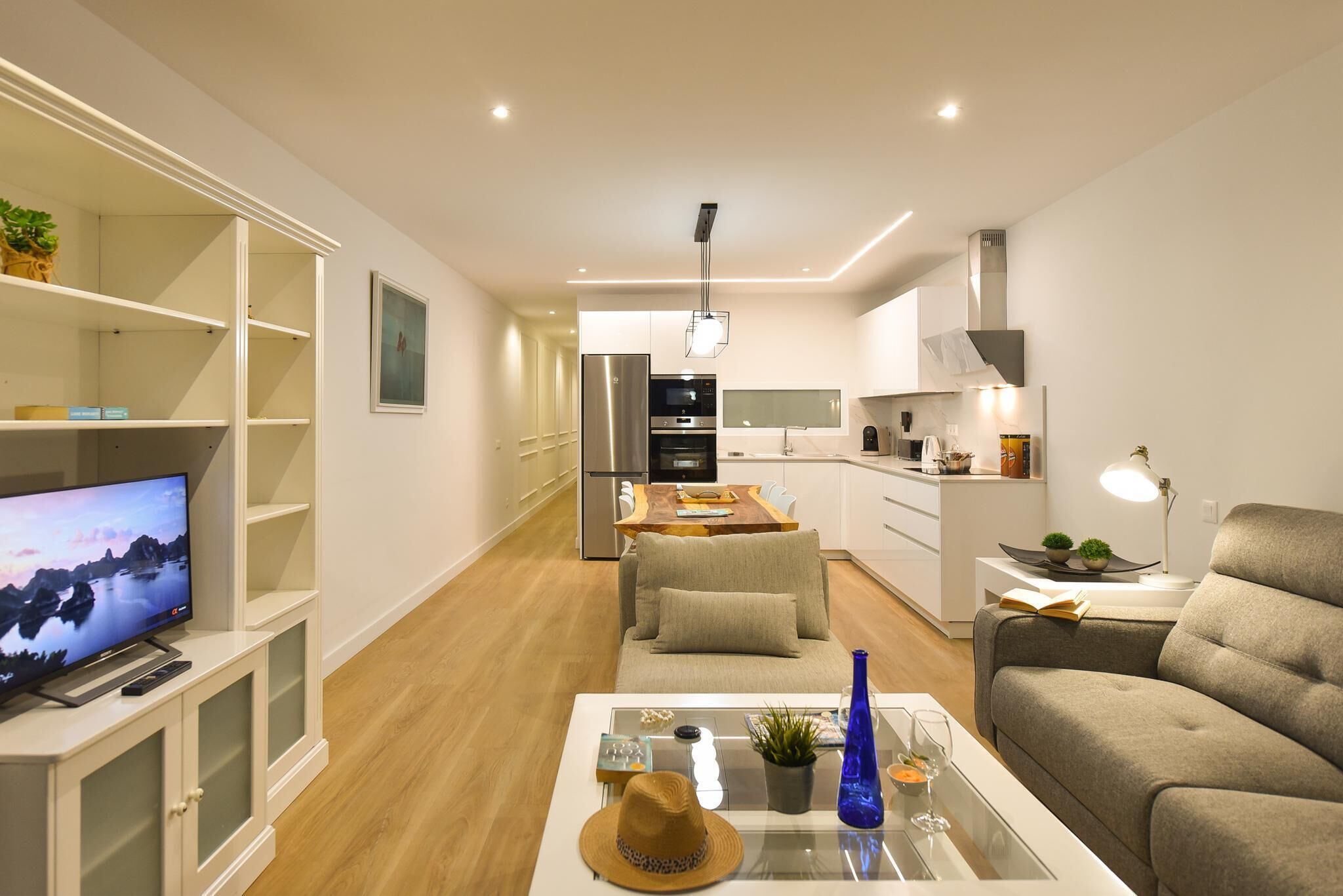 Una sala de estar moderna con televisor, sofá y mesa de centro. Se ve una cocina con comedor al fondo.