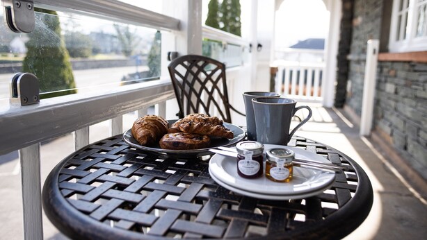 A round metal table with a plate of pastries, a cup, and honey jars on a balcony with a view of trees and a building in the background.