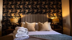 A neatly made bed with a folded white towel, two beige pillows, and a grey headboard. There are two bedside lamps on either side of the bed.