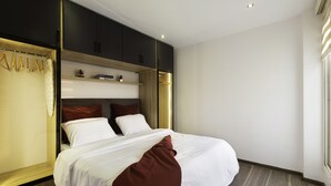 A modern hotel room with a large bed, dark wooden wardrobe, and a small shelf with books.