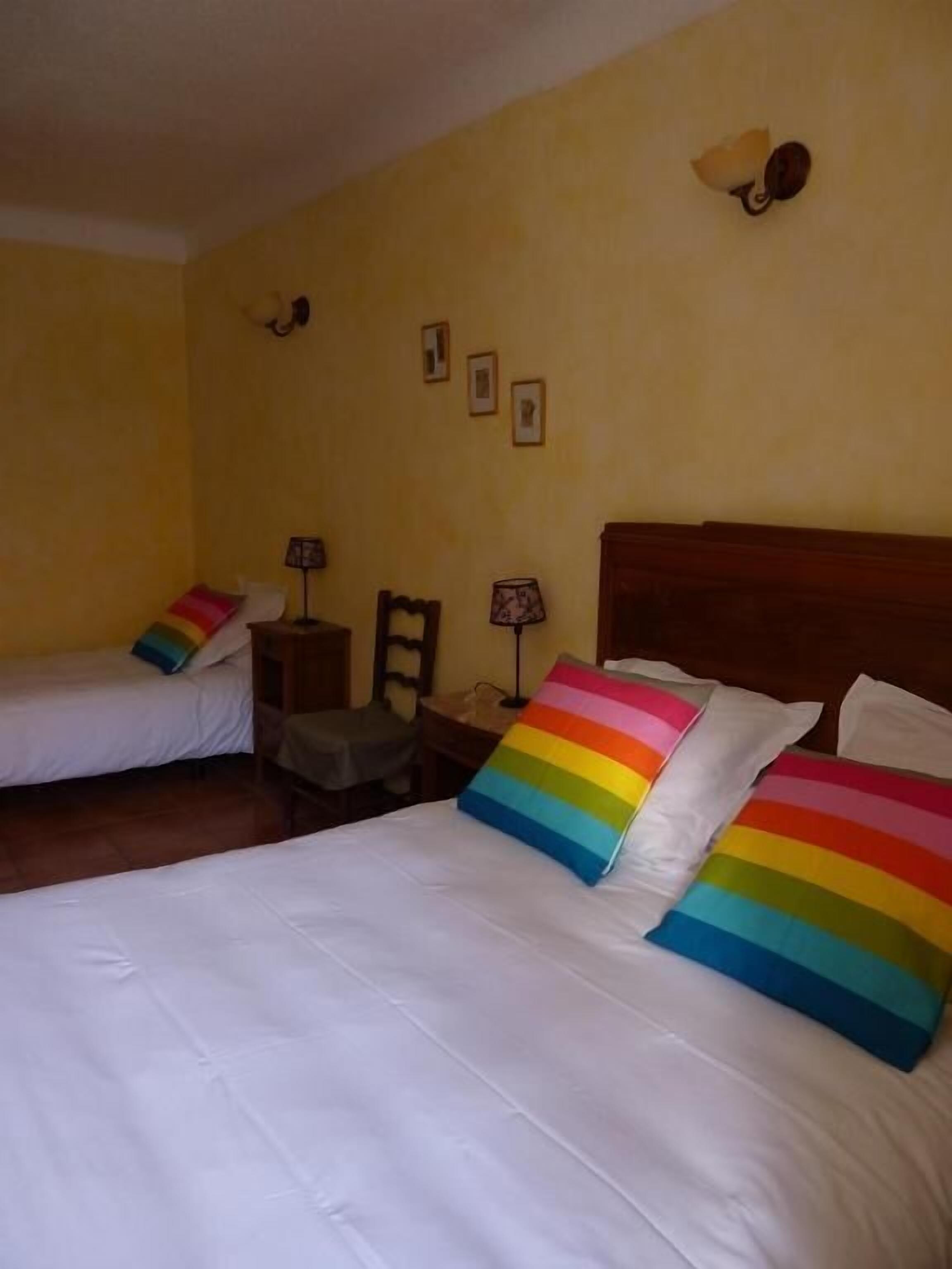 Habitación de hotel con dos camas individuales, cada una con un cojín a rayas coloridas. Hay dos mesitas de noche con lámparas, un cabecero de madera y una silla.
