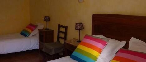 Habitación de hotel con dos camas individuales, cada una con un cojín a rayas coloridas. Hay dos mesitas de noche con lámparas, un cabecero de madera y una silla.