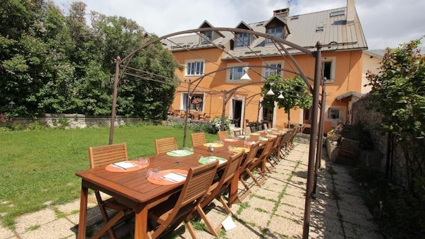 Une longue table en bois, dressée pour un repas en plein air, entourée d’un jardin et d’une maison dont le toit est en pente.