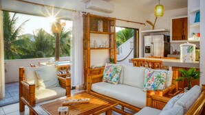 A modern living room with a sofa, chairs, and a coffee table. There is a kitchen area with a refrigerator and cabinets. The room has large windows with a view of palm trees. - DELUXE PENTHOUSE VILLA THE PALMS (El Gariton)