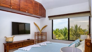 A bedroom with a large bed, a TV mounted on the wall, a wooden cabinet, and a window with a view of palm trees. - DELUXE PENTHOUSE VILLA THE PALMS (El Gariton)