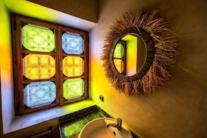 Un baño con una ventana que muestra vidrieras de colores, un espejo redondo con marco de madera natural y un lavamanos.