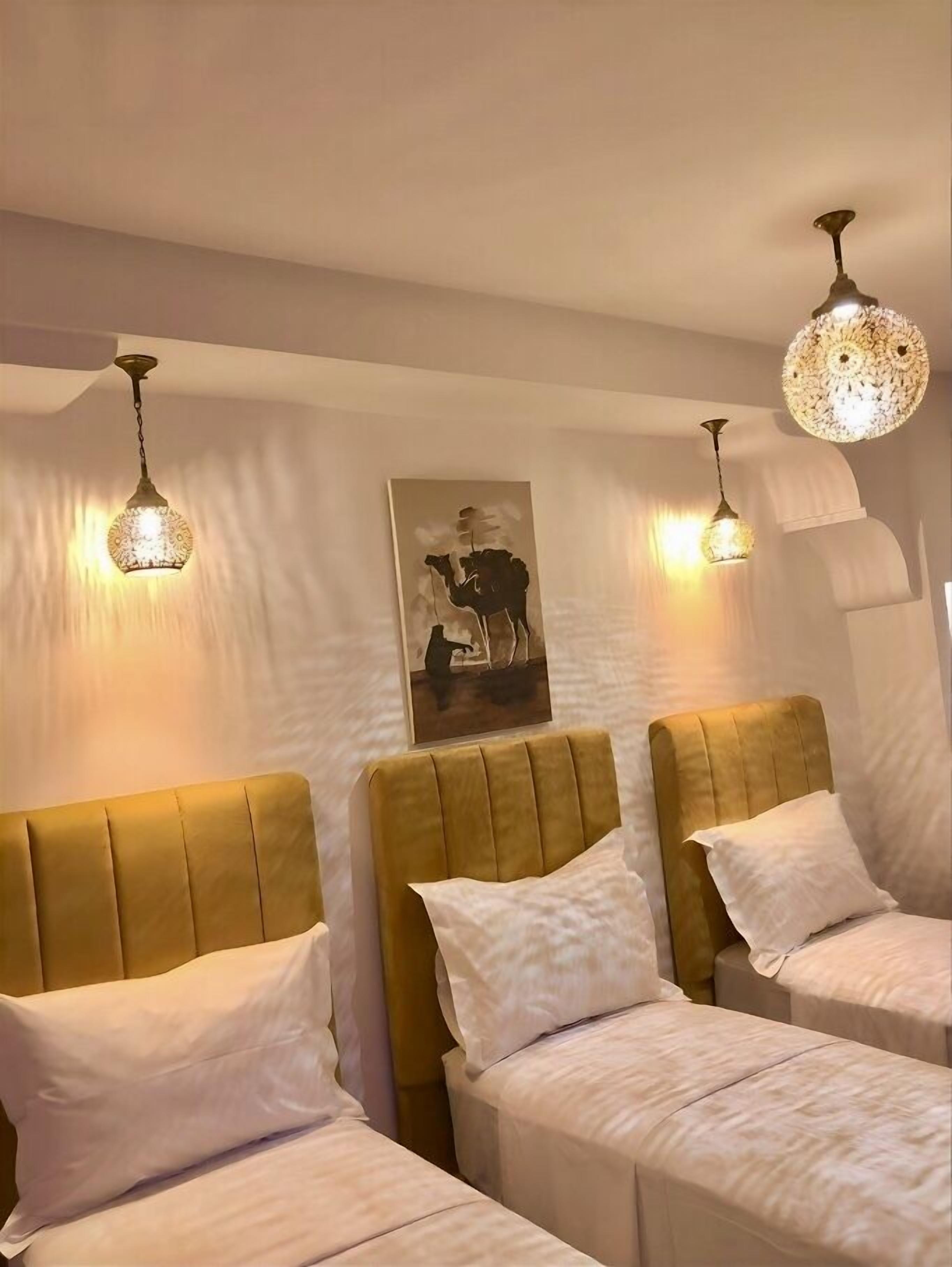 Habitación de hotel con tres camas, cada una con cabecera y almohadas. Se observan apliques de pared y una lámpara en el techo.