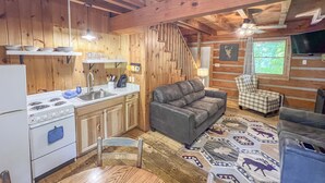L’intérieur d’un chalet rustique, avec des murs en bois, une cuisine équipée d’une cuisinière et d’un évier, un canapé gris et un fauteuil à carreaux.