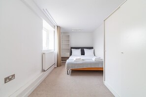 A narrow hotel room with a single bed, a radiator, a window with curtains, and a white door.