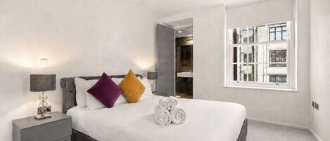 A hotel room with a bed, a nightstand, a lamp, two pillows, a window, and a view of the outside.