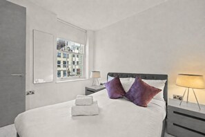A hotel room with a bed, a window with a city view, a bedside table with a lamp, and a nightstand with a lamp. - 1 Min Oxford St: Spacious 2 Bed, Central A/C (London)